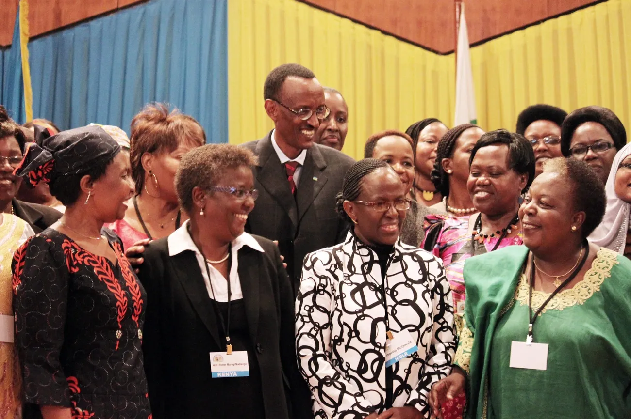 A group of women and men in formal attire stand together in front of a blue and yellow backdrop with a flag.