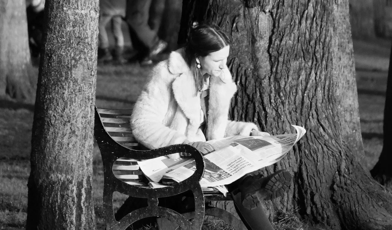Woman in a fur coat sitting on a bench reading a newspaper in a park at night.